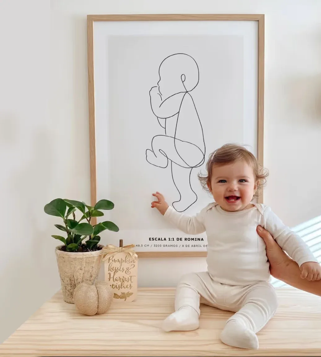 Un bebé sentado en una mesa clara junto a un póster de nacimiento enmarcado con ilustración de líneas y una planta, de la fotografía de Nacío